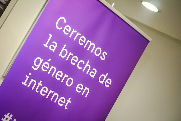 Pendón vertical morado con la frase «Cerremos la brecha de género en internet», iluminado por los focos de la sala.